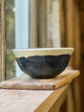 Load image into Gallery viewer, A handmade ceramic bowl with a dark navy blue and black glazed lower half and a light beige unglazed rim, resting on a wooden cutting board on a rustic wooden table near a window.
