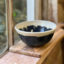 Load image into Gallery viewer, A handmade ceramic bowl with a glossy deep blue lower half and a matte cream upper half, sitting on a wooden coaster on a rustic wooden windowsill.
