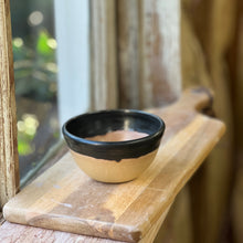 Load image into Gallery viewer, A small handmade ceramic bowl with a black rim and natural beige body, resting on a wooden cutting board on a rustic windowsill.
